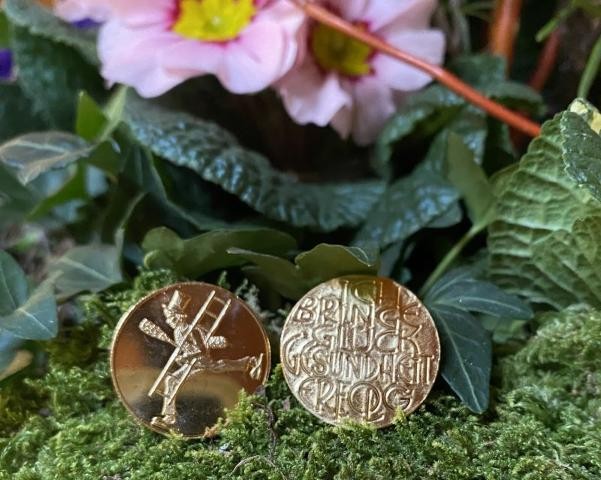 Zwei goldene Glücksmünzen mit Schornsteinfeger vor rosa Primel im Moos